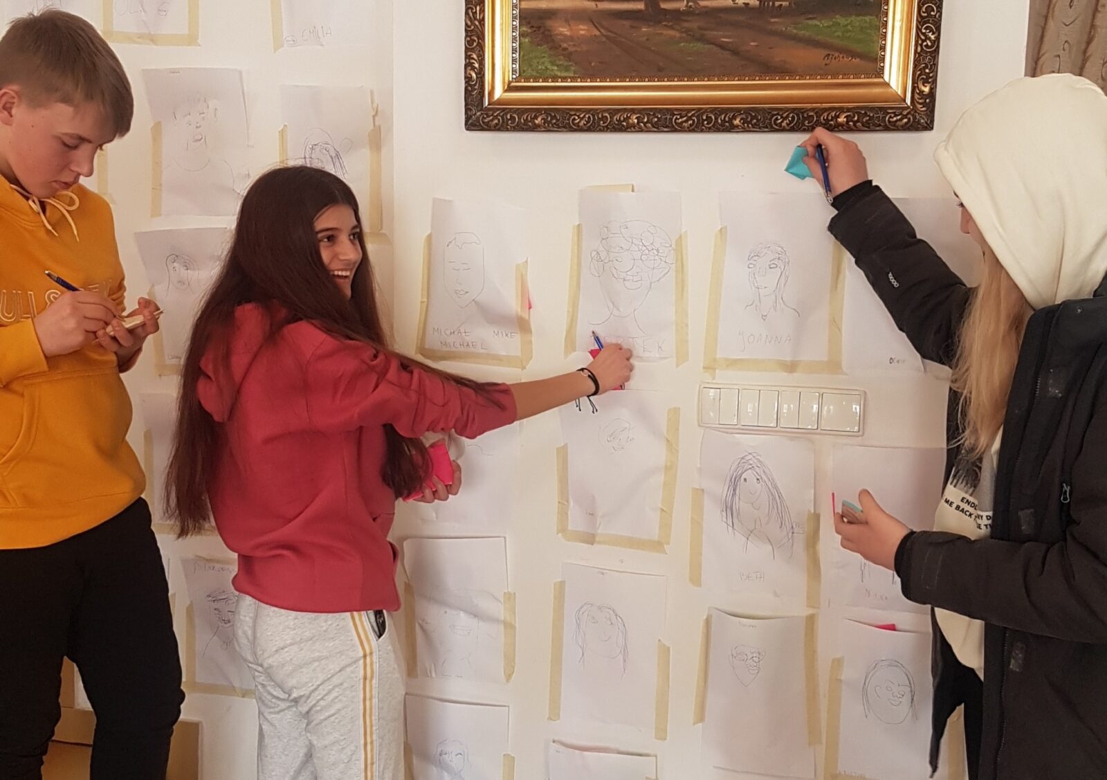 Tre ungdommer skriver på postit-lapper foran en vegg med artige portrettbilder