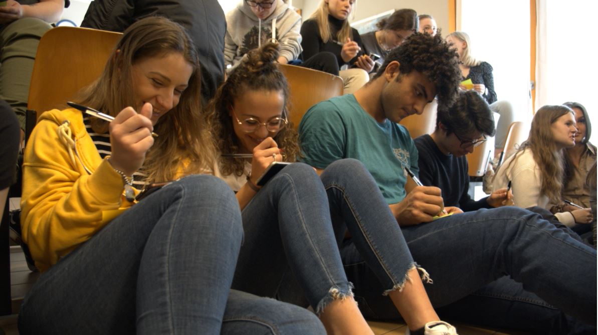 Ungdommer sitter på rekke og rad og smiler mens de skriver på lapper