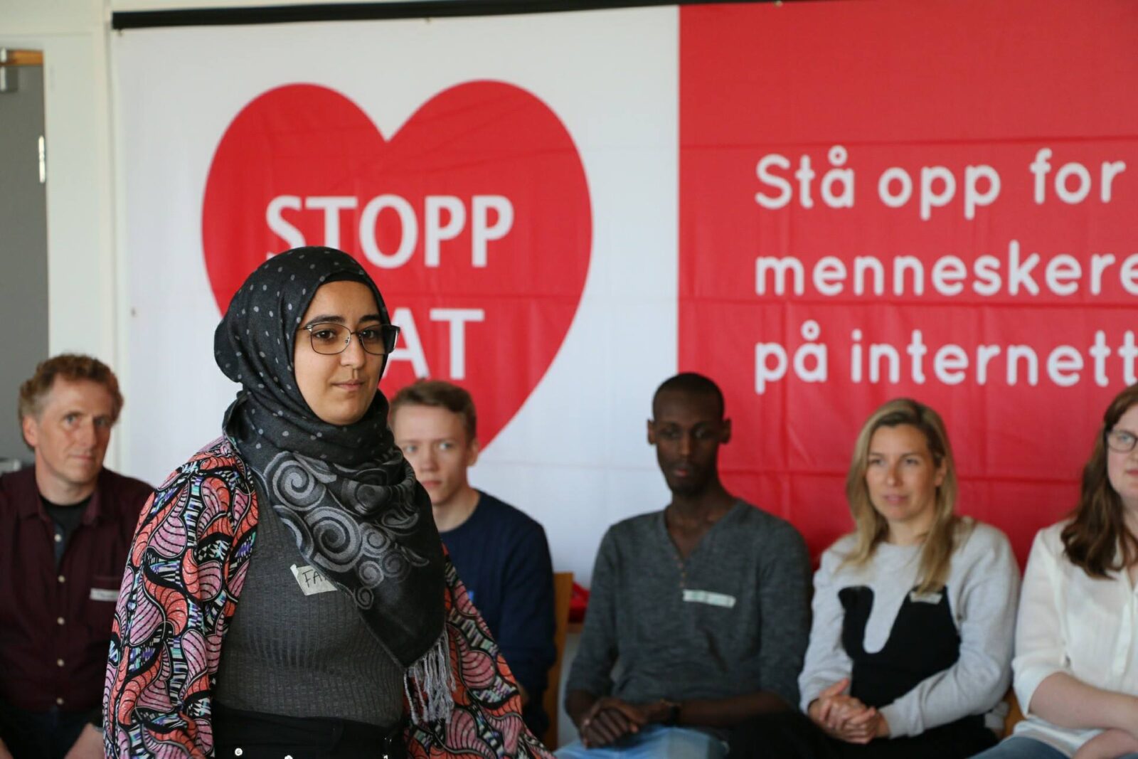 En ung kvinne kvinne går forbi deltakere som sitter langs en vegg med en stor plakat i bakgrunnen hvor det står Stopp hat