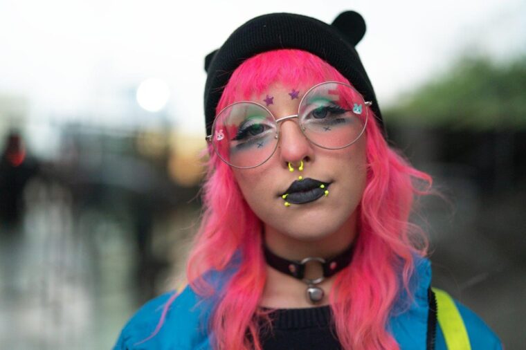 En ung kvinne med rosa hår, store briller, svart leppestift og gule piercinger
