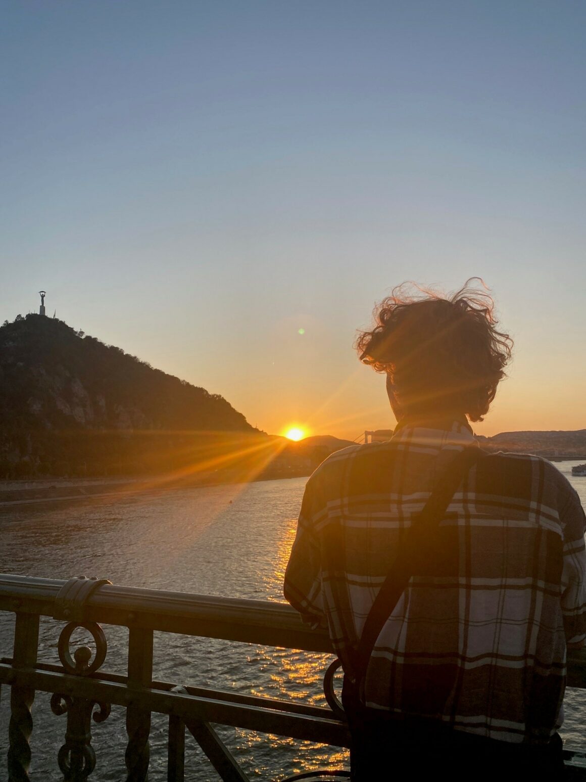 En ung gutt står med ryggen til kamera på en bro og ser på en solnedgang.