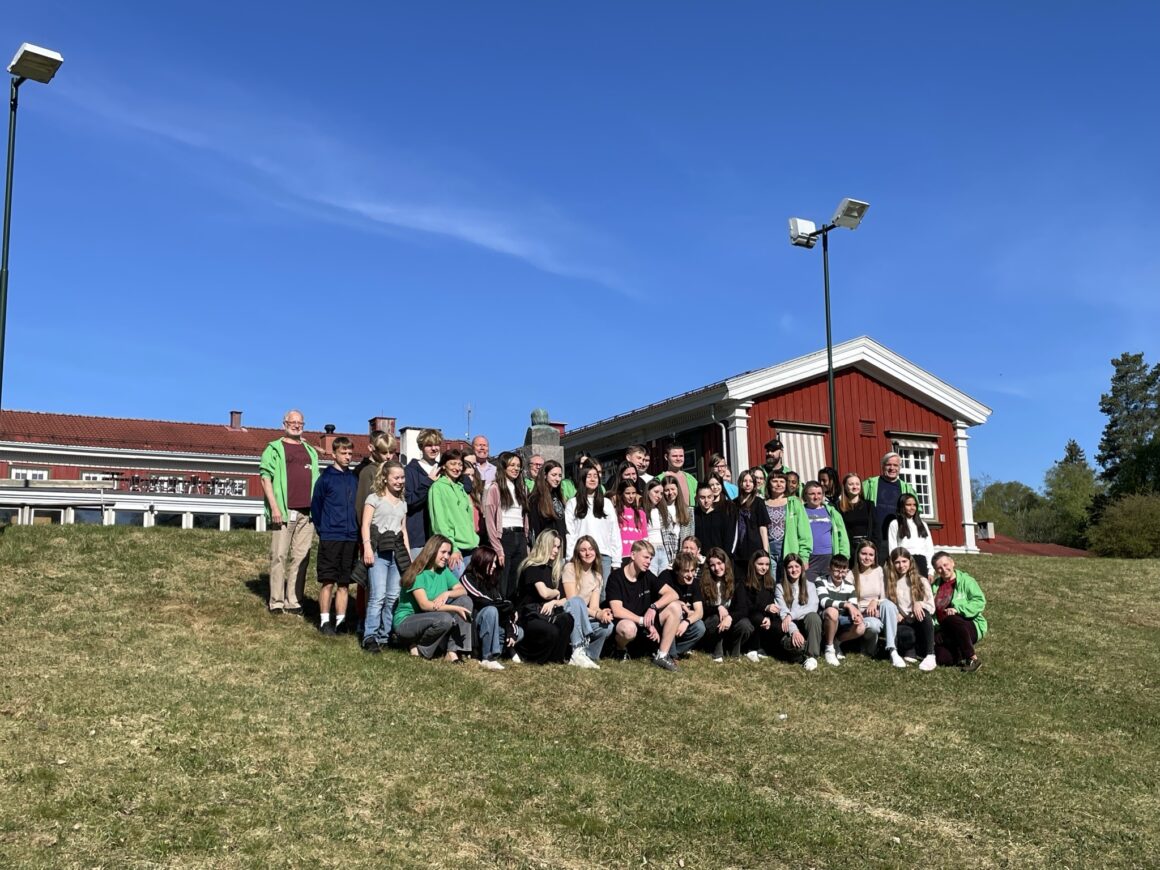 En gruppe ungdommer står oppsamlet for et gruppebilde foran et rødt hus. Sola skinner.